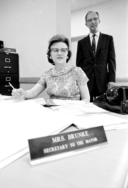 Adeline Brunke is shown at her desk in the Mayor's office on her last day before retirement. She served as secretary for four Madison mayors in the past seven years. Her final boss, Mayor Otto Festge, is standing at right.