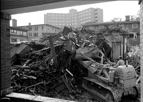 Workers and equipment are shown tearing down old student dwellings to make room for the Elvehjem Art Center complex. It is surrounded by Park Street, University Avenue, State Street, and Murray Street. The view is looking toward Park Street and Chadbourne Hall.