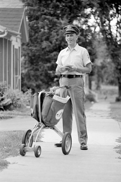 John L. Smith standing on a Madison sidewalk with letters in his hand and his mail cart beside him. He is featured in a newspaper article about his busy life. Besides his job he is a father, church leader, resident manager of an apartment house, and sports enthusiast.