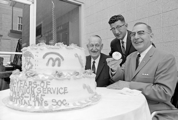 Three men posing behind a cake. One of them is holding up a cupcake displaying a traditional credit union symbol. Left to right are the decorated commemorative cake; Charles Hyland, member of the 1935 CUNA Mutual board of directors and retired comptroller; E.C. Davis, center, president of the Wichita (Kans.) postal employes credit union; and Charles F. Eikel Jr., holding a cupcake displaying a traditional credit union symbol.