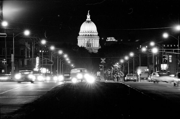 The Wisconsin State Capitol at night from West Washington Avenue. There is a neon or electric sign for "Hotel Loraine." 