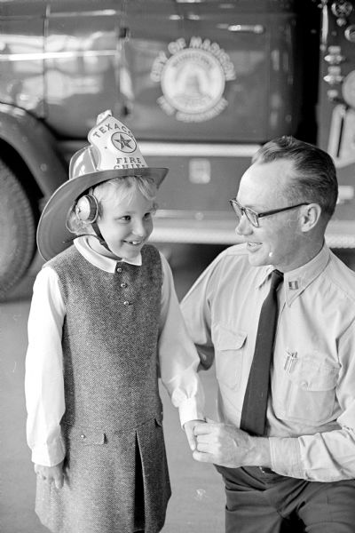 Ann Peterson, 9, of 2111 Clyde Gallagher Dr., proudly dons the loudspeaker-equipped fire helmet she won after visiting No. 3 fire station, 1217 Williamson St., during Fire Prevention Week.  Capt. Charles Hessling is looking on. A fire truck is parked behind them.
