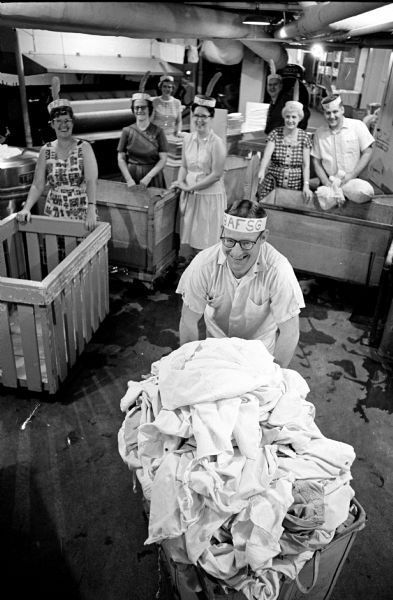 Slightly elevated view of Laundry Crew members wearing head bands at work in celebration of being the first unit in Madison to give 100% of its quota as Fair Share Givers. In front is laundry manager William Peterson. In the background, left to right: Ethel Jasper, Peggy Lawton, Lea Peterson, Loretta Taggert, Wesley Josephson, Pearl Rodefeld, and John Baukin.