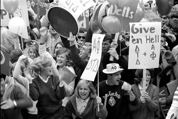 A crowd of Delta Tau Delta and Alpha Phi members are shown cheering at a Badger Homecoming Rally. Some of the people are carrying signs.