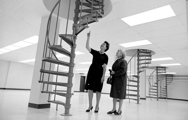 Mary Albers, assistant cashier at Security State Bank, is shown pointing upward to show Martha Horstmeyer the new spiral staircases built to enable bank tellers to climb up to the drive-in banking cages.

A second image is a photograph of an ad for Security State Bank's public tour of their newly remodeled facility. This photo was taken earlier (October 16, 1965).