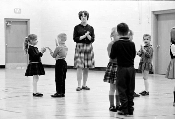 Thirteen lay teacher's assistants are hired to assist teachers in nine different Madison schools where the number of students exceeds 30 or 32 in the classroom. Lay assistant Kathy Tiltbach is shown teaching numbers to students using a clapping routine.