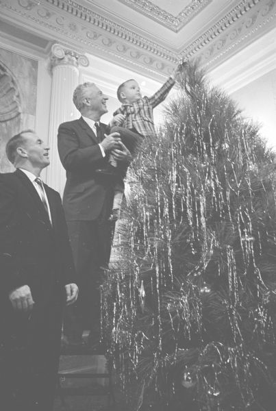 Four-year-old John Steigerwaldt is putting a decoration on the top of the Christmas tree in the governor's office. He is being held up by Governor Warren Knowles. At left is Richard Jeffery, Oconomowoc, area manager of the General Tree Corporation. John is the son of Edward Steigerwaldt, Tomahawk, president of the Wisconsin Christmas Tree Producers Association.