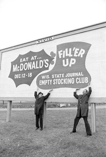 Drawing attention to a large billboard sign is Edwin Traisman, left, president of McDonald's of Madison, and Vernon Cox of Vivid Outdoor Advertising Company. Words on the signs read: "Eat at McDonald's Dec. 12 - 18" and "Fill'er Up, Wisconsin State Journal Empty Stocking Club." The sign is located at 125 Fair Oaks Avenue. Other billboard sites with this sign are at 2201 South Park Street, 3317 University Avenue, and 3051 East Washington Avenue.