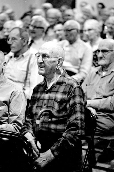 An unnamed "Dane oldster," a resident of the Dane county home, wearing a plaid shirt and holding a cane, is sitting in the audience and watching unseen entertainers from the Kehl dance studio. The event is the annual Christmas party sponsored by the Madison Assn. of Life Underwriters.