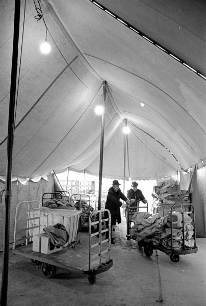 Two postal workers arranging mail carts full of "yule mail" inside of a large canvas tent. The men working are George DeVoe, left, a superintendent engineer, and Leonard Roe, a general mechanic. The tent was a wind buffer over the loading area at the temporary post office at the U.S. Army Reserve Center at 1402 S. Park street. 