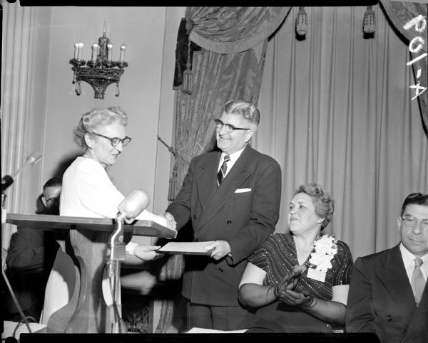A man is presenting an award to a woman at a podium.  Another woman, wearing a corsage, is looking on and clapping.