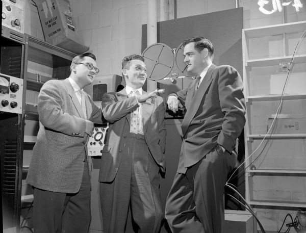 Three men are standing in the WMTV studio. There is a film reel in the background.