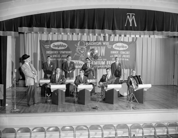 Uncle Julius and his 8 piece band on a stage with a banner in the background that reads: "Hear the Doughboy, WKOW Breakfast Symphony with Uncle Julius, 6 - 6:15 am, Dinner Concert with Uncle Julius 12 - 12:15 pm."