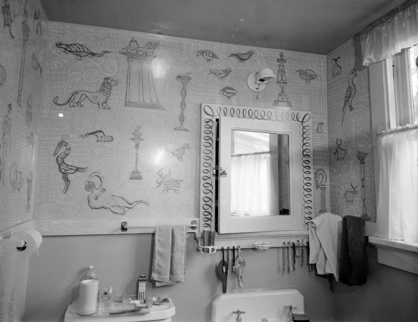 Interior of Aaron Bohrod's bathroom, 715 E. Gorham Street. His drawings decorate the upper walls of the room, including the mirror frame. An assortment of toiletries are arranged on the toilet tank. An assortment of hairbrushes and toothbrushes are hanging under the mirror, including a child's toothbrush in the shape of a gun in a holster.