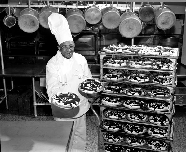 Chef Carson Gulley holding two pies and standing in front of a rack of fudge bottom pies.  He is posing for a WMTV ad for his cooking show to be broadcast on the new TV station WMTV. Full biographical sketch of Mr. Gulley can be found in the associated newspaper article.<p>The recipe can be found in Gulley, Carson "Seasoning secrets and favorite recipes of Carson Gulley" Copyright 1956.<p>A digital version can be found on the University of Wisconsin-Madison Libraries, Digital Collections, The Human Ecology Collection, Gulley, Carson / Seasoning secrets and favorite recipes of Carson Gulley (1956) Page 106.</p>