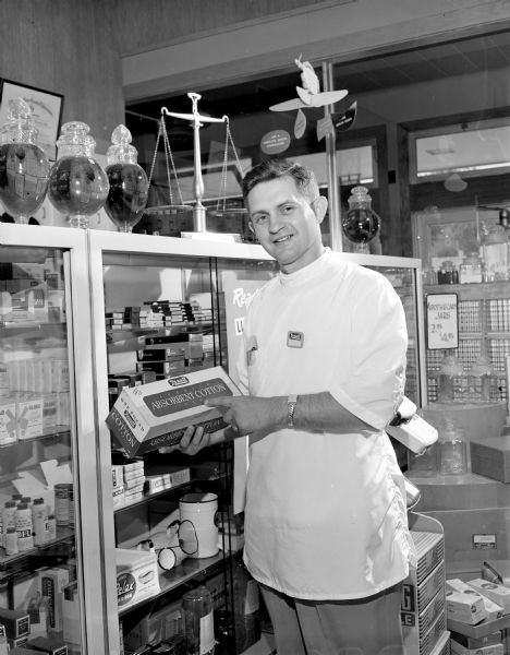 Druggist holding a box of Rexall Absorbent Cotton and standing in front of a display case at Rennebohm's Drug Store #15 in the Shorewood Corners Shopping Center, 3250 University Avenue.