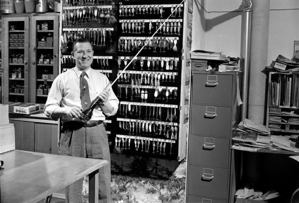 Alvin Lewison holding a fishing rod and standing in front of a rack of fishing lures in his Sportsmen's Home sporting goods store in the Shorewood Corners Shopping Center, 3240 University Avenue.