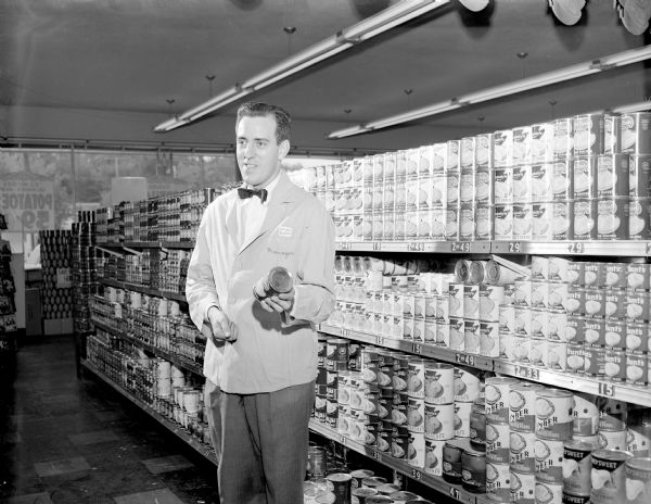 Salesman holding a can of food standing in front of shelves holding cans of food in the National Food Store, Shorewood Corners Shopping Center, 3244 University Avenue.