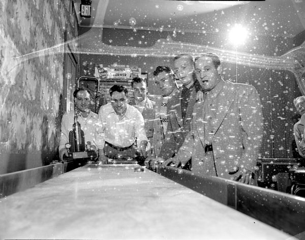 Six men are standing behind a shuffle board table, with one man holding a trophy, in John Gervasi's Tavern, 713 Regent Street.