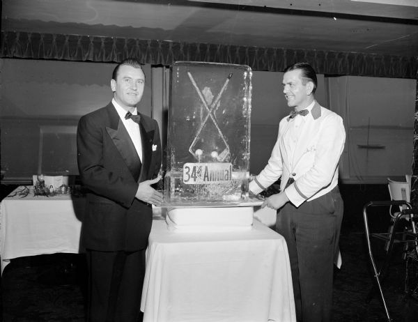 Two men in formal dress standing beside a block of ice which is imbedded with two small golf clubs and two golf balls and a sign that reads: "34th Annual".