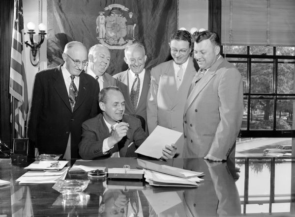 Five men standing behind Gov. Walter Kohler watching him sign a document in his office. One of the men might be Paul Thielan from Brookfield, Wisconsin.