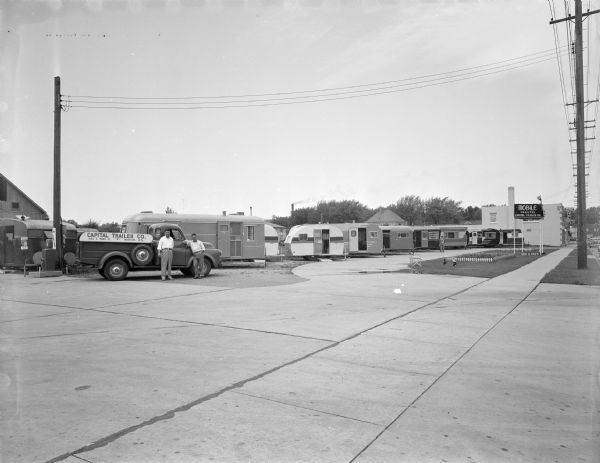 Two men standing beside a truck with a sign reading: "Capital Trailer Co. 960 S. Park St." There are trailers in the background.