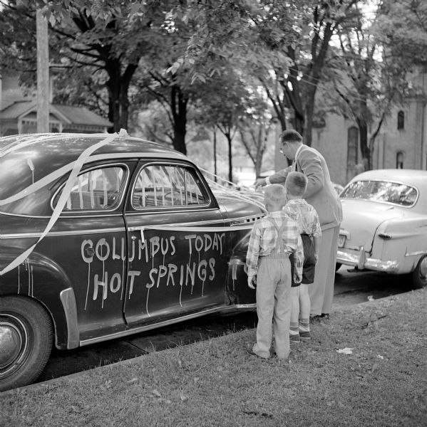 View towards a man and two boys decorating the "get-away" car for the wedding of Dave Wilson and Jean Goodell. The sign sprayed on the side of the car reads: "Columbus today, Hot Springs [Tomorrow]".