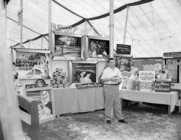 View towards a man standing in the Miller Brewing Co. exhibit booth under a tent, sponsored by the East Side Liquor Store, 1047 Atwood Avenue and distributed by Simon Brothers Co. Inc., at the East Side Business Men's Association Festival. The exhibit includes four framed pictures of people enjoying life at these venues: golf, baseball, pool and beach. Signs read: "Enjoy Life with Miller High Life" and "Guess how many bottle caps are in the jug" .