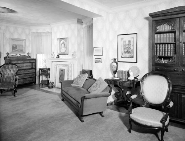 Living room in the Vaughan house. There is fireplace with a framed portrait of a woman above the mantel, and a framed portrait of a man propped on the floor over the front of the fireplace.