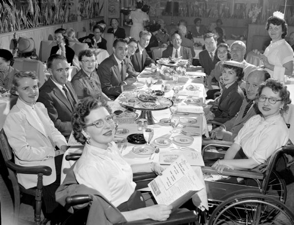 Fourteen men and women (one in a wheelchair) sitting around a dining table in a restaurant (Hoffman House?) as part of the WKOW-TV telethon (?).