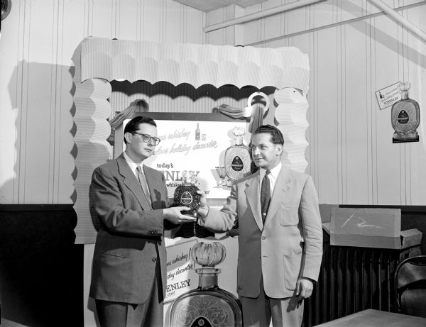 Two General Beverage employees holding a bottle of Schenley whiskey between them and standing in front of a Schenley Whiskey display booth.