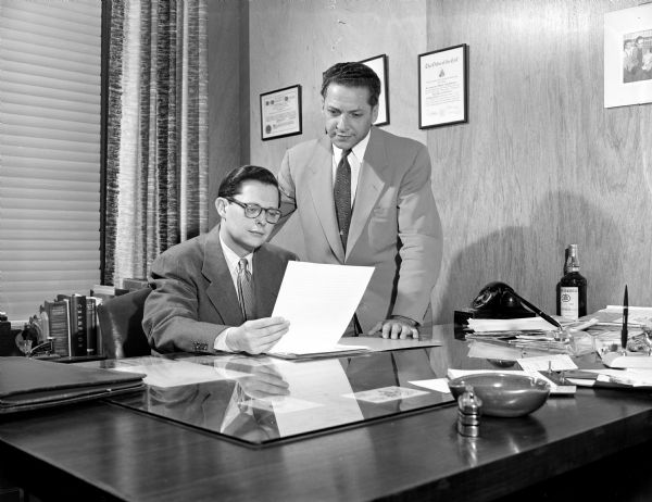 Two General Beverage Sales Co. employees. One man is sitting behind a desk, and the other man is looking over his shoulder.