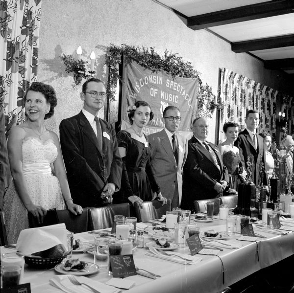 Drum Corps leaders and chaperones at the head table of the banquet honoring the midwestern champion Boy Scout Drum and Bugle Corps at the Nakoma Country Club. From left to right: Mrs. and Mr. Henry Johnson, Mrs. and Mr. Roger Muzzy, Clarence H. Beebe, Maryon Mesenkamp, and Stan Stitgen. The cloth banner on the wall reads, "Wisconsin Spectacle of Music."