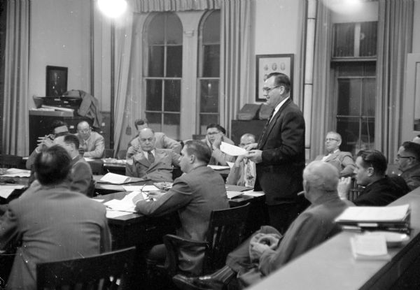 Spectators and members at the Madison City Council meeting on November 24, 1953.  Mayor George Forster (standing) has the floor. Aldermen shown to the left of Forster, counter clockwise, are Archie Simonson, George Reger, A. William Johnson and Ivan Nestingen.