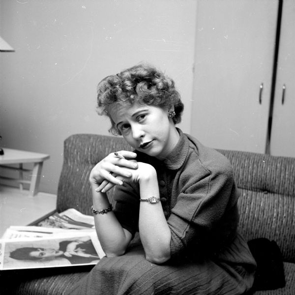Portrait of Mrs. Edwin (Donna) Stein. She is sitting on a couch next to portrait prints of herself, and is holding a cigarette. She is wearing a wristwatch and a bracelet. 