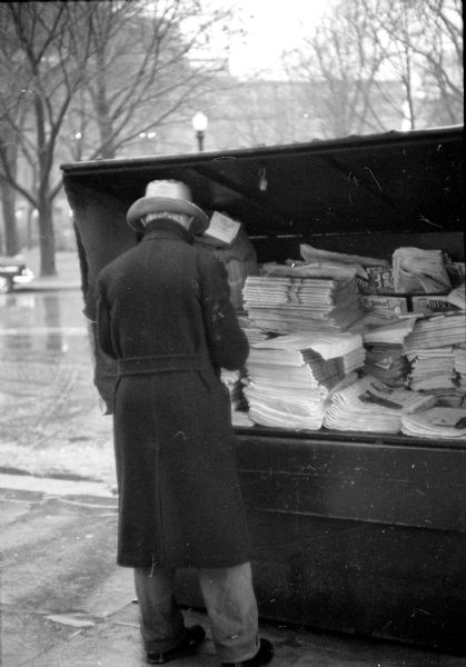 A newspaper salesman guarding his wares against a damp chilly wind. 