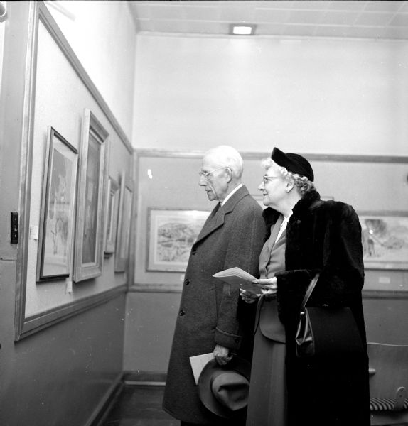 William and Louise Hussey compare their program with one of the exhibited framed paintings at Madison Art Association Collectors' Show in Scanlan Hall at the Madison Vocational School.