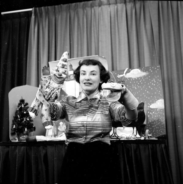 Merrillyn (Mrs. Robert) Wegner is shown holding puppets and standing in front of a set from "Your Puppet Pals", a weekly show on WKOW-TV. She has a smiling clown, in a conical hat and waving, on her right hand, and a dragon with mouth agape on her left. There is a miniature Christmas tree and backdrop of clouds and snowy sky on the desk in the background. Merrillyn was known in later life as Lynn Hartridge.