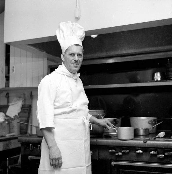 A chef working in a large kitchen. He is wearing a white chef's coat, tall hat and crisply pressed apron, and is standing next to an industrial-sized stove with his hand on the handle of a metal pot. Next to the pot is a large metal spoon. Several other sizable sauce pans and stock pots are at the back of the stove. 