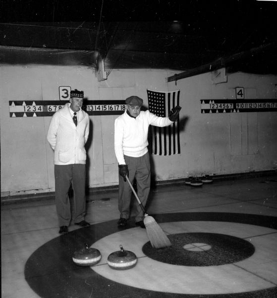 Two members of the Madison Curling Club during an intra-club competition. Norm Sonju (left) and Gil Bach survey shots made by members of their rinks. Bach, looking down at the floor of four concentric circles, is holding a broom in his hand, the end of which is lying to the side of two curling stones. Sonju is looking up with a cigarette in his mouth, and his hands behind his back. They are each wearing white (or cream) colored sweaters, gloves, slacks, and distinctive hats. An American flag is hanging on the wall behind them between score cards.