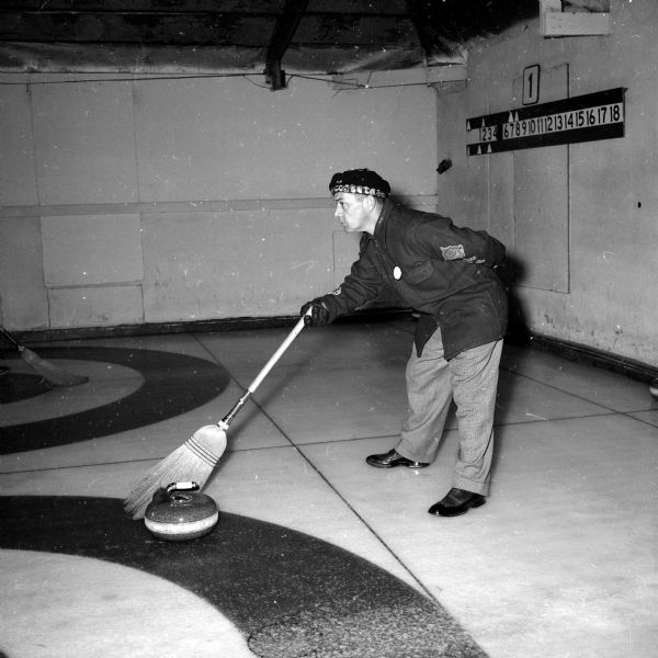 A member of the Madison Curling Club during an intra-club competition. Gloved curler, Jim Cottrell, is standing on the outer circle, pointing with his broom in the direction he wants a member of his rink to "lay" his stone. 