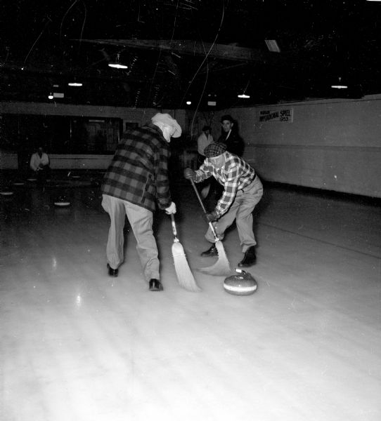 Two members of the Madison Curling Club during an intra-club competition. Gloved curlers, L.W. Ketchum (left) and Rollie Black (right), show a "sweeping" technique with brooms as an attempt to coax a rink mate's stone. 