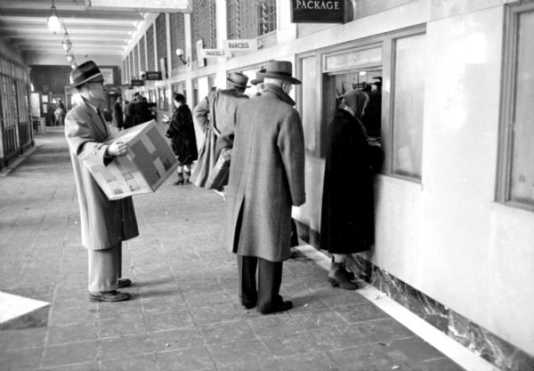 People waiting in line to mail their letters, packages and parcels in the lobby of the United States Post Office on Monona Avenue. A woman is standing at the window, making a transaction, while two gentleman are lined up behind her, one with a large square box in his hands. In the background, other customers are at other service windows. 