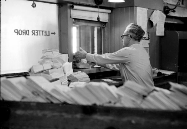 Postal employee, Harry Bunbury, receiving bundles of Christmas cards from inside the letter window at the United States Post Office on Monona Avenue. Bundles of sealed envelopes are stacked in the foreground and near his desk. 