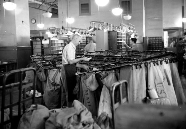 Postal worker, Del Winters, working at a pouch rack, where mail is sorted prior to going out of the city. He is in the back of the United States Post Office on Monona Avenue with other co-workers who sort letters.