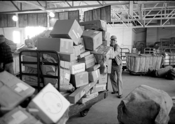 Postal worker, Ray Kindschi, is pushing a load of packages on a cart at the Williamson Street warehouse for the United States Post Office. He is usually a mail truck driver but now is assigned to routing incoming packages, given the volume at the holiday time of year.