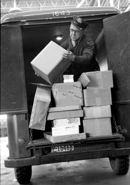 Postal worker, Paul McConnell, loading packages onto his mail truck for delivery. He is handling different sized boxes stacked on the lowered gate of his open truck.