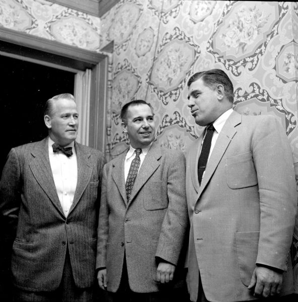 Three members of the Rounders Club meeting at the annual Christmas party held in the Heidelberg Hofbrau. Milt Bruhn, University of Wisconsin line coach (right) is talking with Vern Woodward (left), university assistant boxing coach, and Warren Lamm. They are standing in front of a wallpapered wall, just inside a framed doorway. 