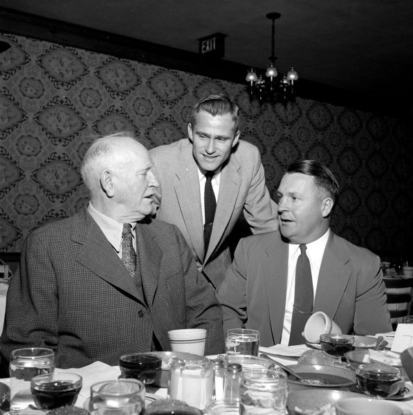 Three members of the Rounders Club meeting at the annual Christmas party held in the Heidelberg Hofbrau. In the dining room sitting at a table, Lew Burdette (center), ace pitcher for the Milwaukee Braves who spoke to the club, is listening to William McCormick (left), retired chief of the Madison Police Department. Hugh Reynolds (right) is also listening.  