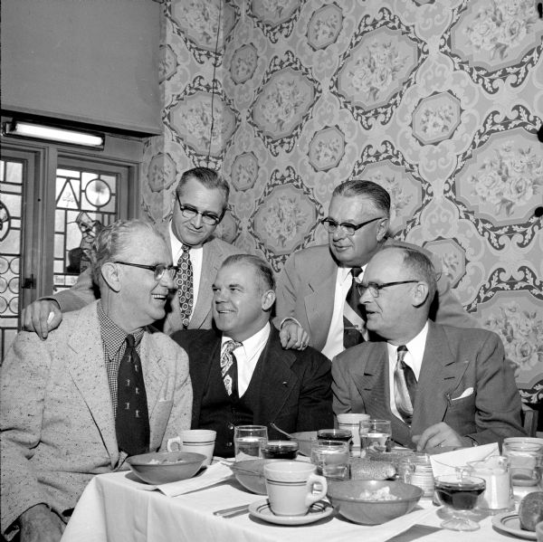 Five members of the Rounders Club meet at the annual Christmas party in the Heidelberg Hofbrau. Roundy Coughlin, eternal president of the Rounders Club, gives the word to Olaf Severson, the treasurer (seated center), and Al Larson, secretary (seated right). Listening are Ray Messner (rear left) and Bob Holmes. They are sitting just inside a dining room with elaborate floral wallpaper and leaded glass window panels. 